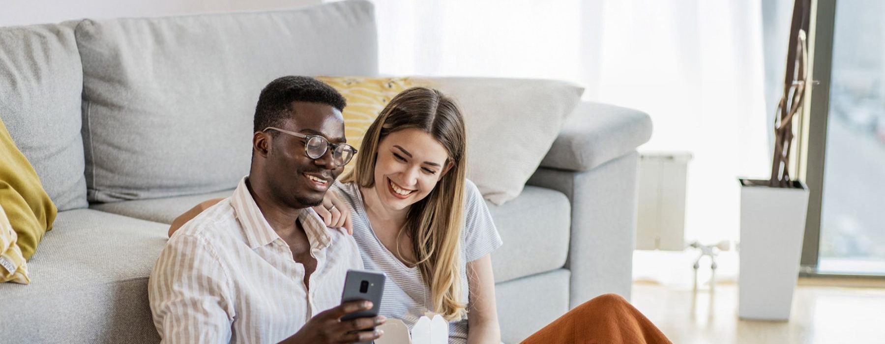 A man and woman with take out, sit against a couch on their living room floor and watch at their cell phone